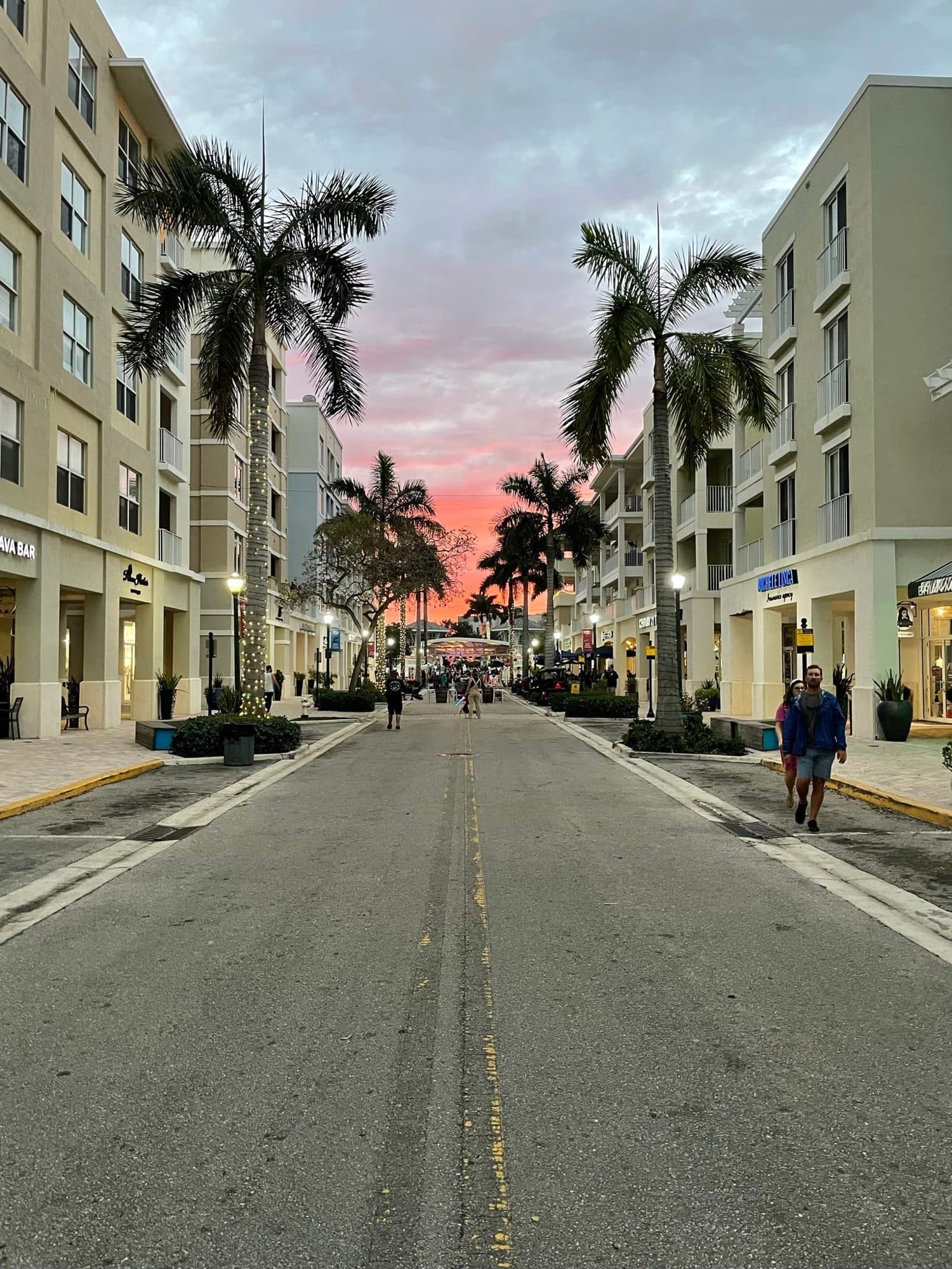 Palm-lined main street in Abacoa Town Center at sunset, with shops and apartments on both sides and a few people walking