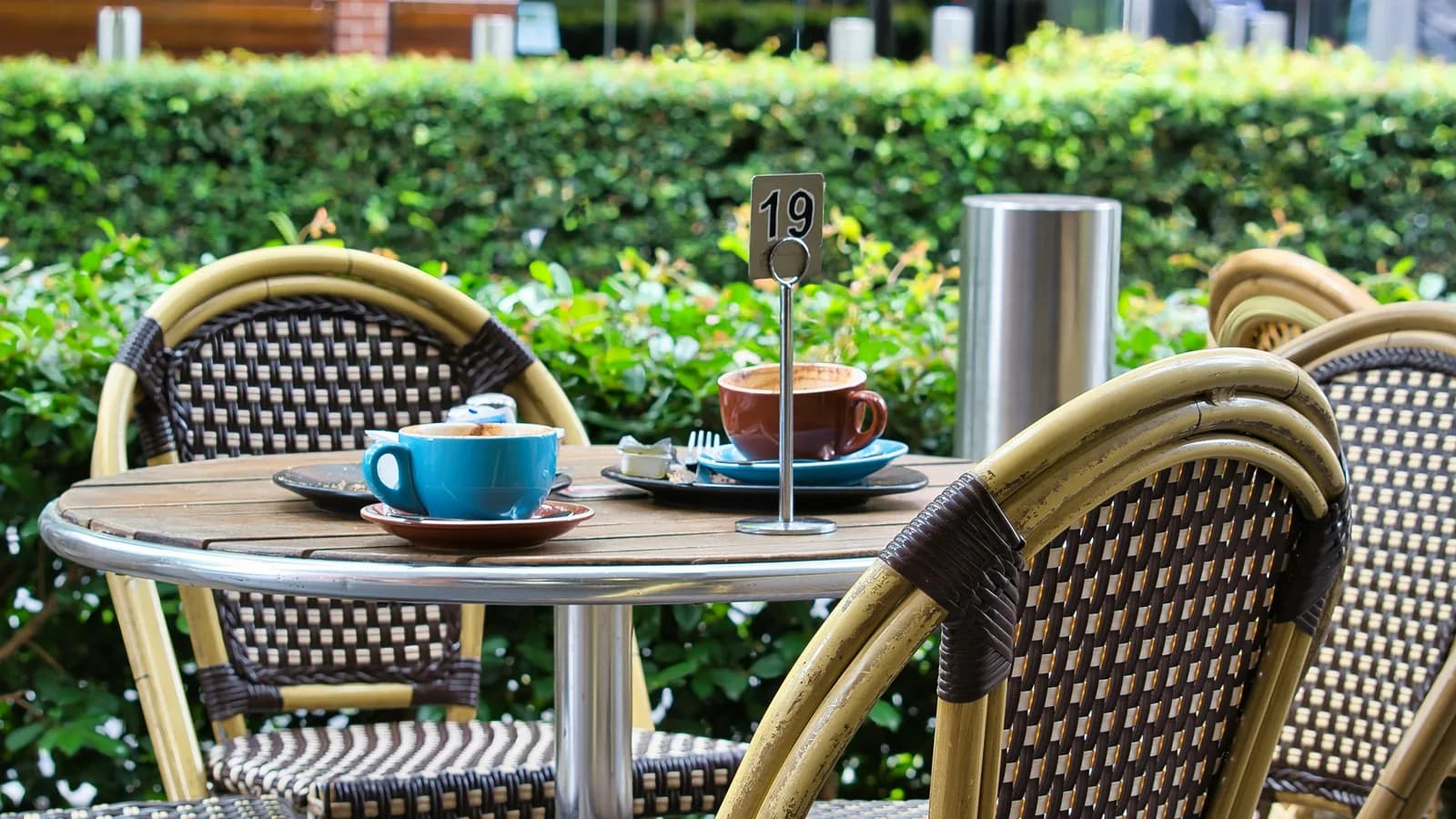 An outdoor café table with two cups of coffee, rattan bistro chairs and a clipped green hedge behind