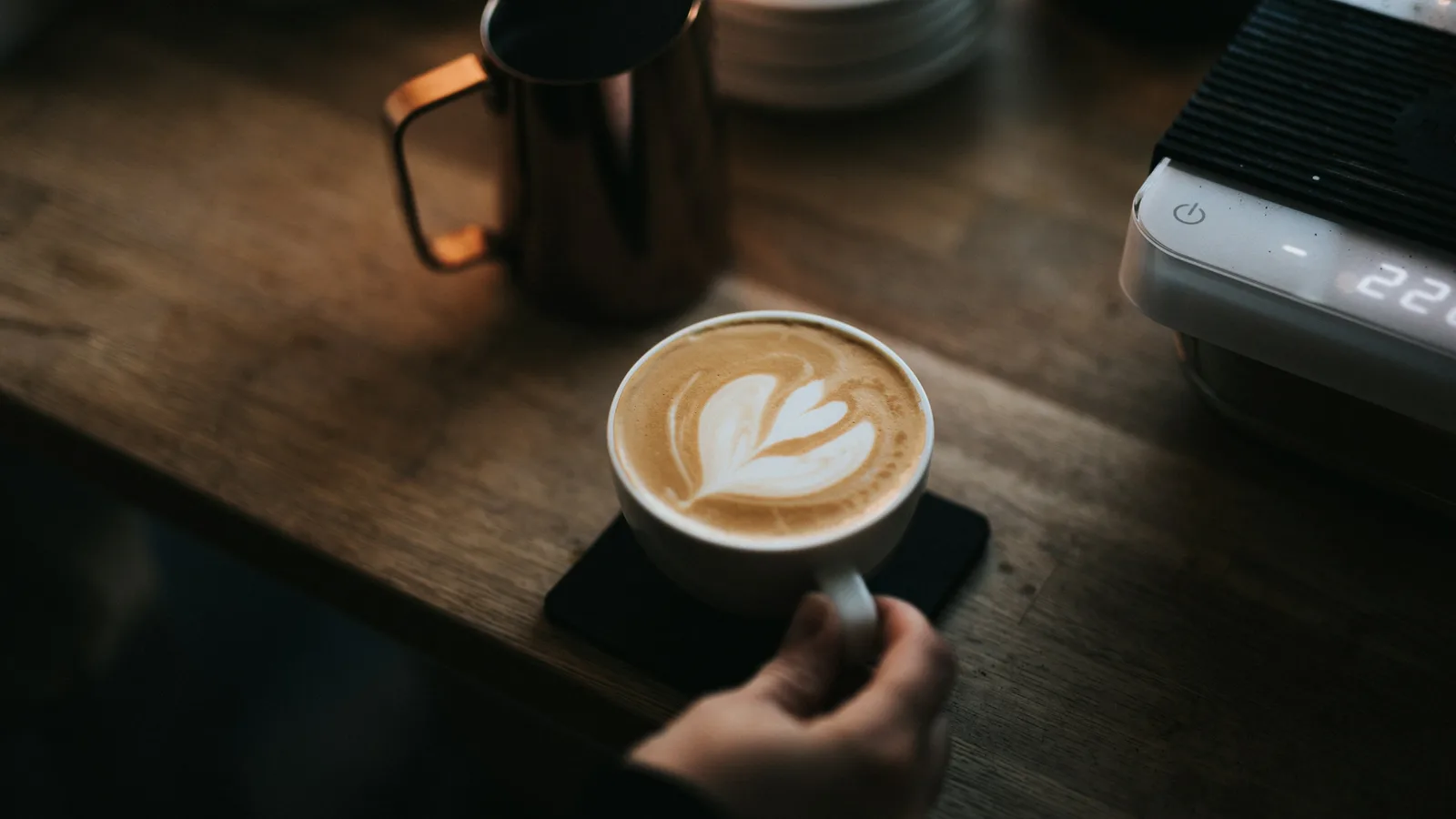 A cappuccino with latte art in a white cup on a dark wooden café counter, beside a milk jug and an espresso scale