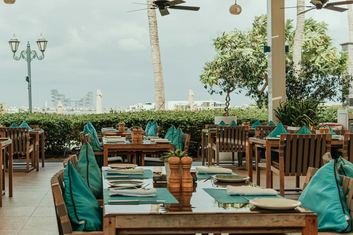 A covered open-air dining terrace with wooden tables set for service, teal napkins and cushions, ceiling fans overhead and palms beyond the hedge
