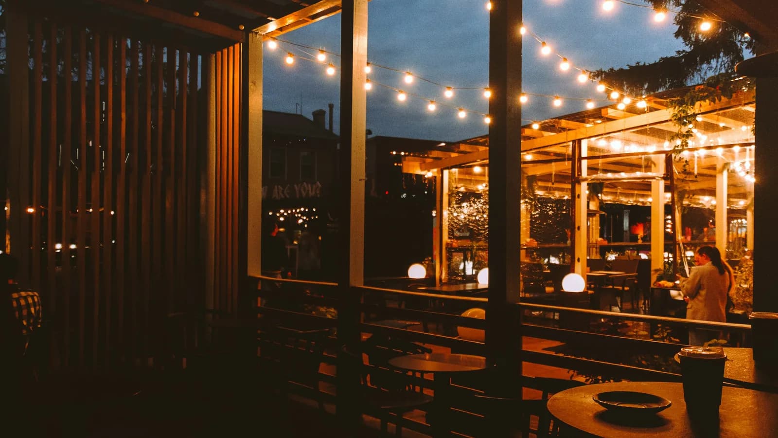 A covered patio at dusk strung with warm bulb lights, with empty tables in the foreground and a lit dining area beyond