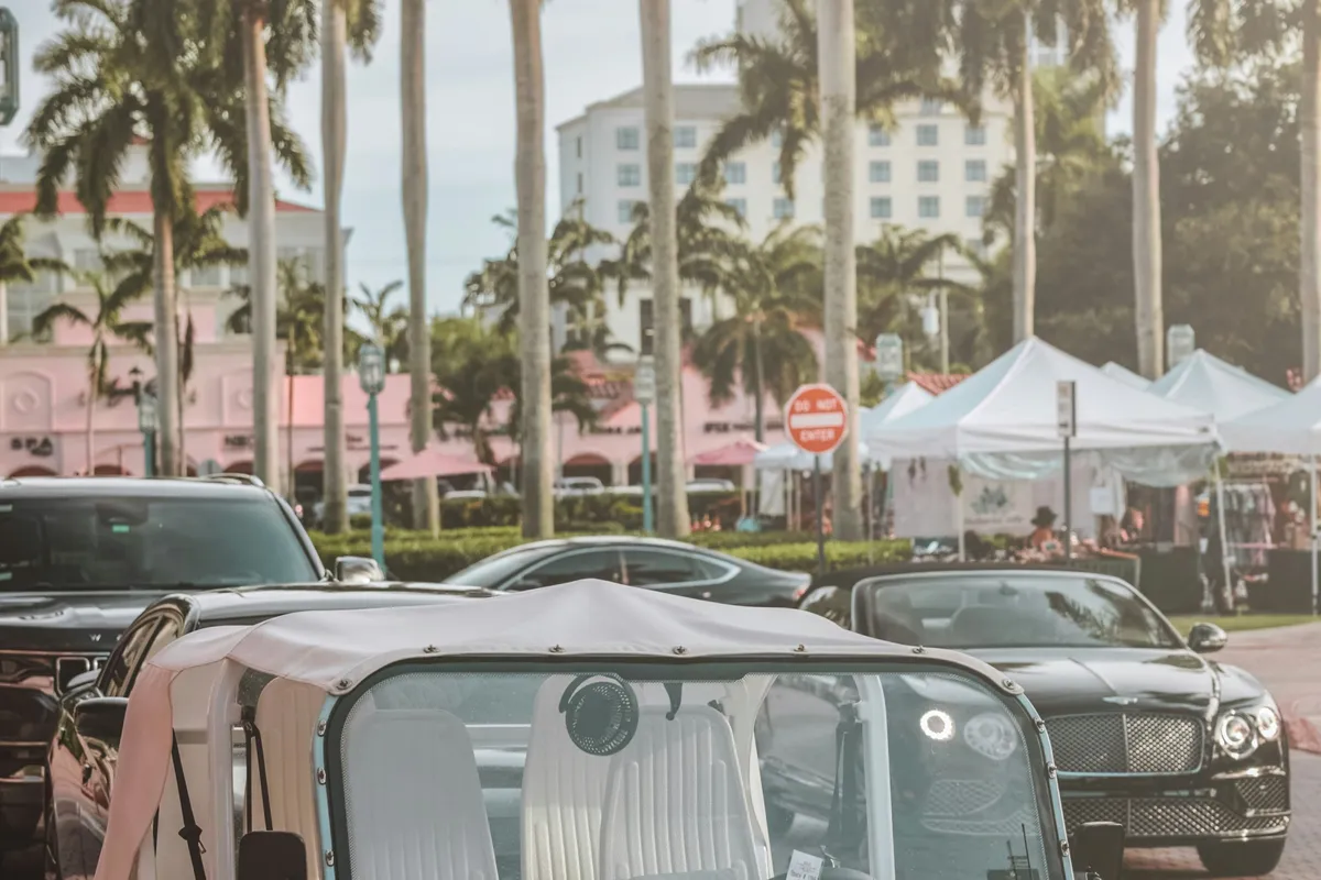 Royal palms above pastel Mediterranean-style storefronts and white market tents, with the canopy of an open-top Moke in the foreground