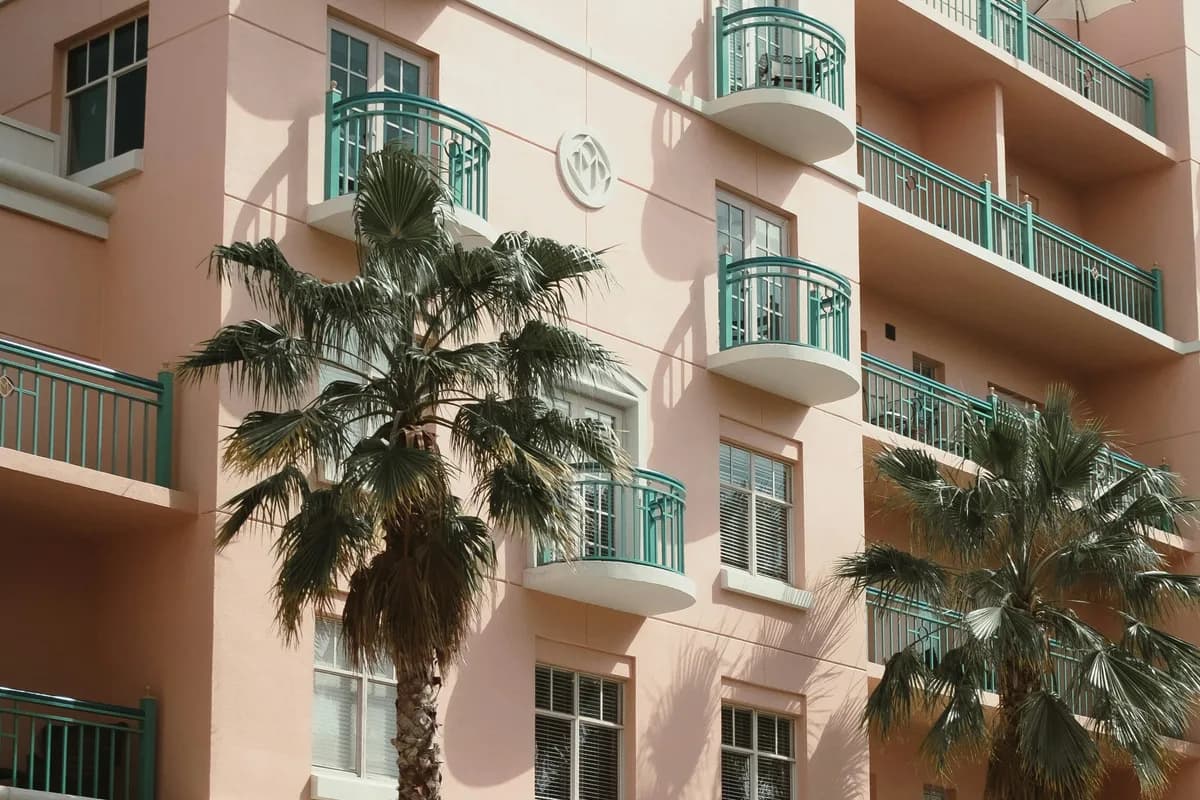 Upper floors of a pastel Mediterranean-style building with teal balcony railings, a patio umbrella, and palm trees against a partly cloudy sky