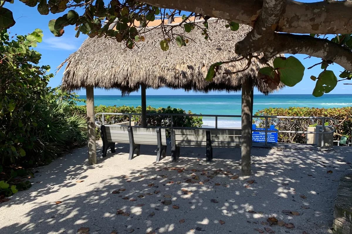 A thatched chickee with benches at a beach access, shaded by sea grape branches, looking out over a railing to turquoise ocean, with a Palm Beach County parks sign at right