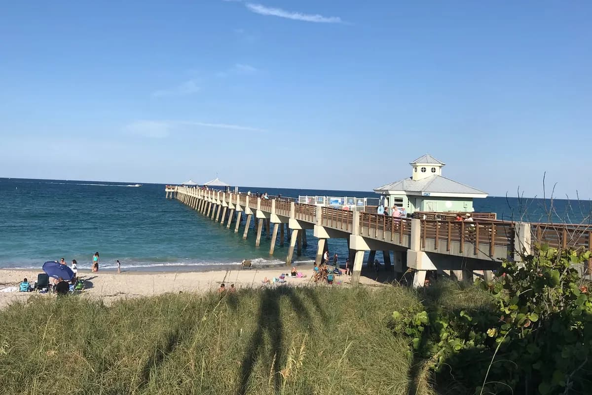 The Juno Beach Pier reaching into the Atlantic beside its pale green pier house, with beachgoers on the sand and sea oats and sea grape on the dune in the foreground