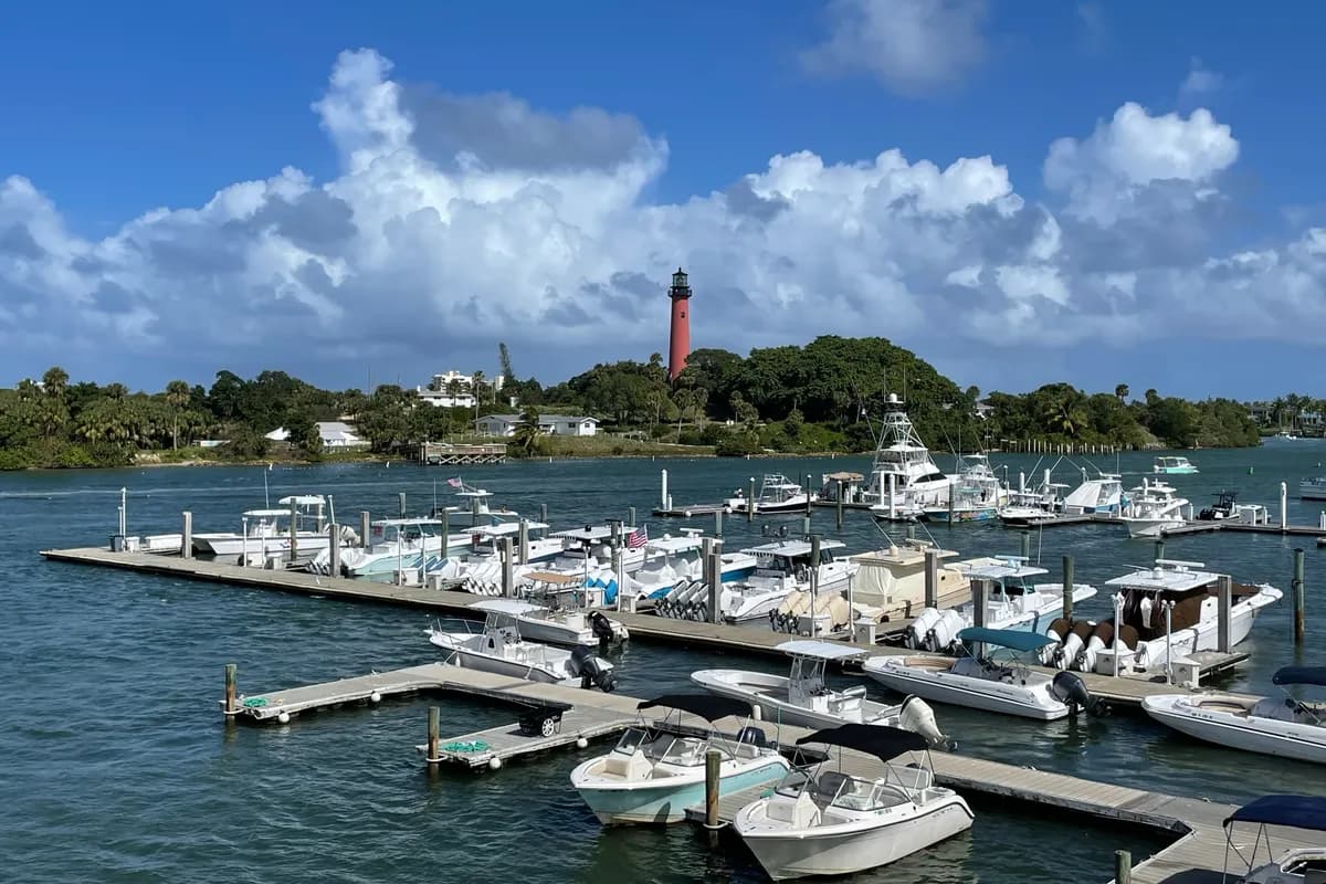 The red Jupiter Inlet Lighthouse in Jupiter, on a wooded rise across the water from a marina full of boats, under big white clouds