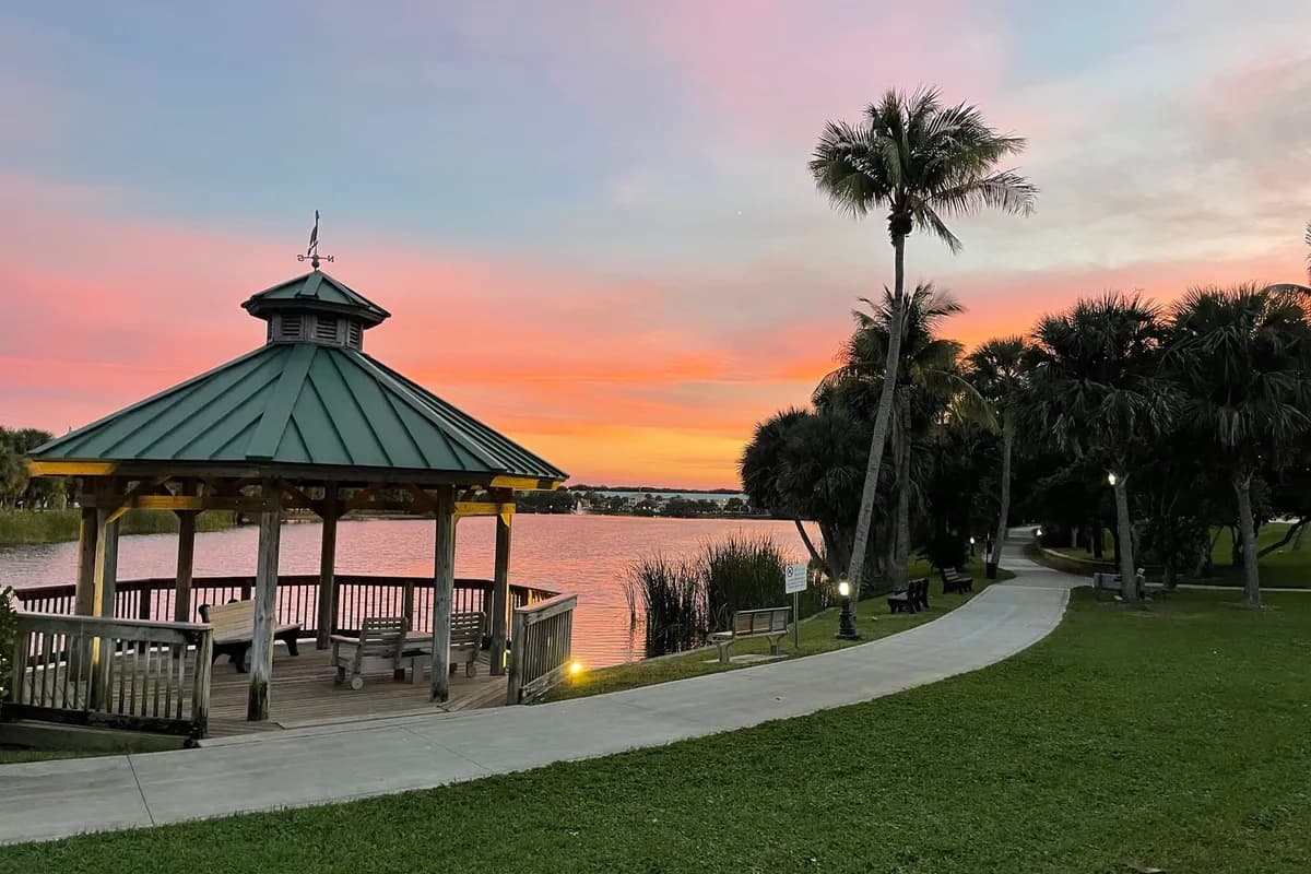 A wooden gazebo with a green metal roof beside Pelican Lake at sunset, with a curving path, benches, lamp posts and palms