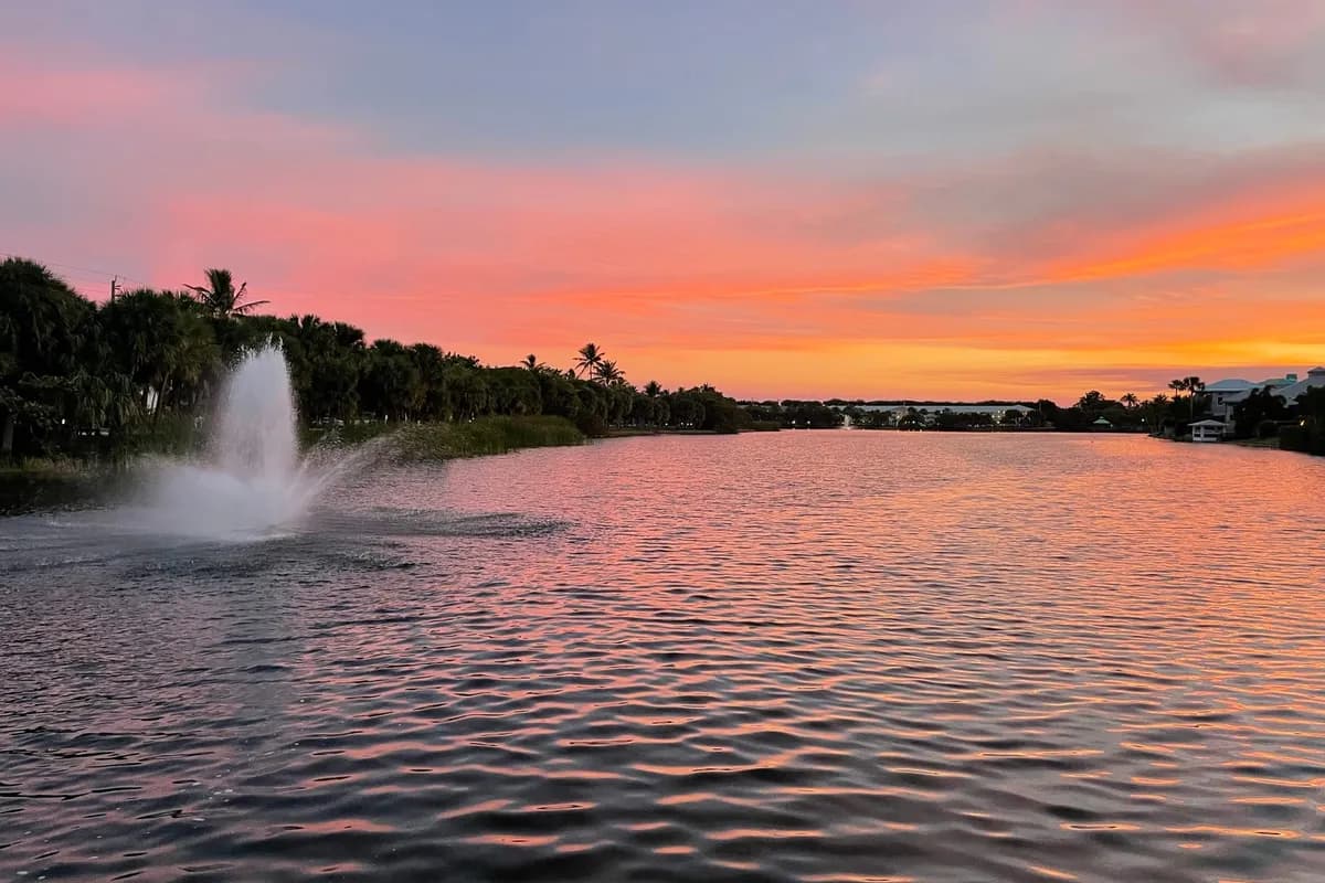 Pelican Lake in Juno Beach at sunset under an orange and pink sky, with a fountain at left and homes along the far shore