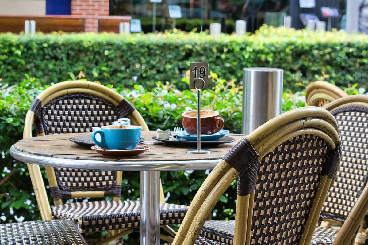 An outdoor café table with two coffee cups, finished breakfast plates and a table-number stand, between rattan bistro chairs in front of a green hedge