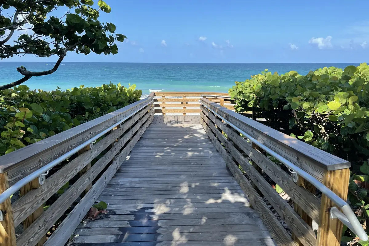 A wooden dune crossover boardwalk with metal handrails leading between sea grape hedges toward a turquoise ocean under a clear blue sky in Jupiter