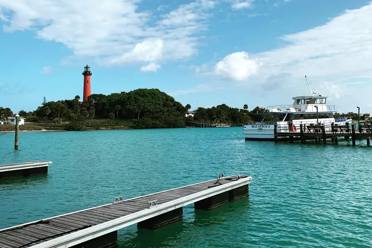 The red Jupiter Inlet Lighthouse above trees on the far shore, a white two-deck tour boat tied to a wooden pier at right, and a floating dock in the foreground on turquoise water under a blue sky with clouds