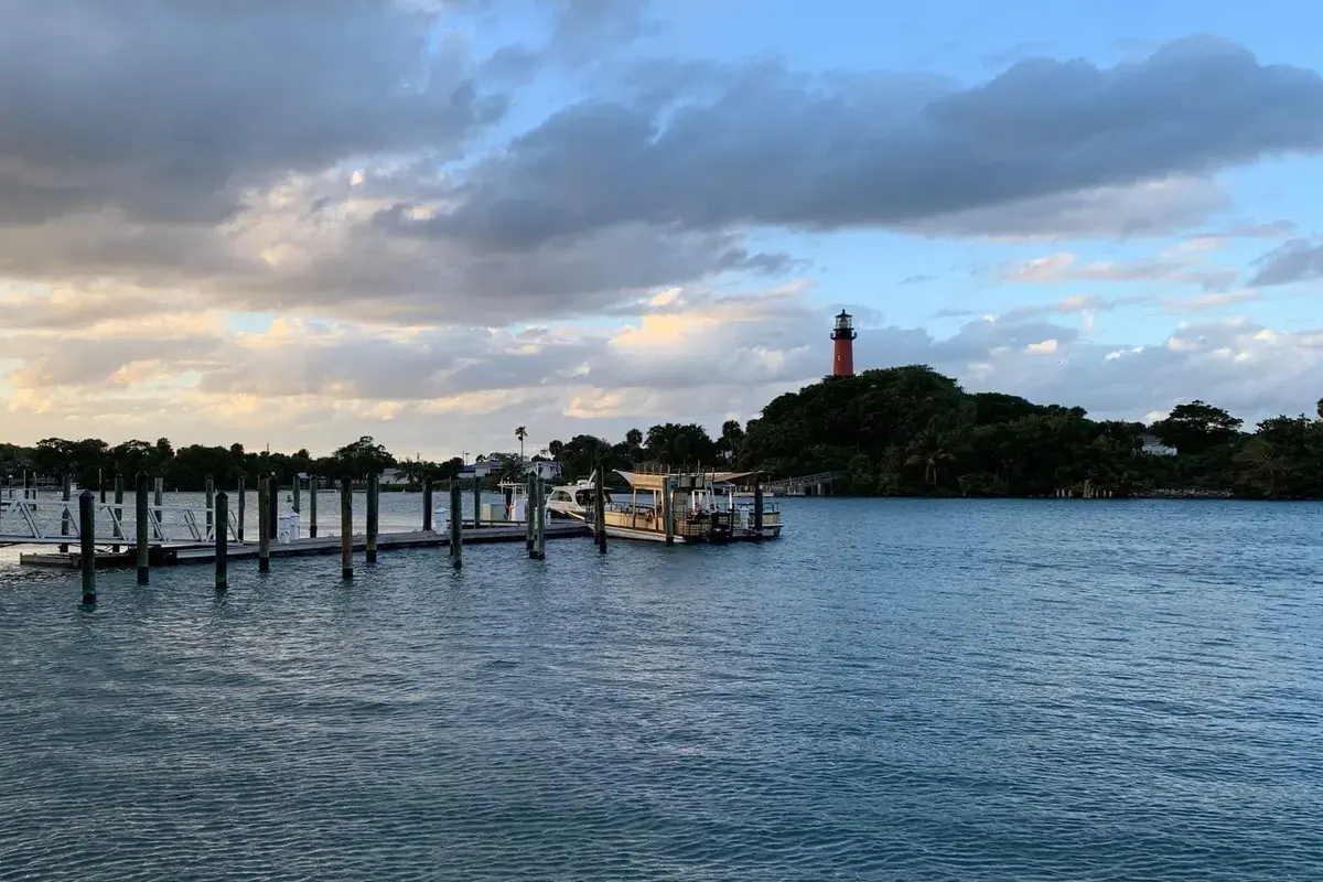 The red Jupiter Inlet Lighthouse on its wooded point at dusk under a bank of clouds, seen across the inlet past a line of dock pilings and a floating dock in the foreground