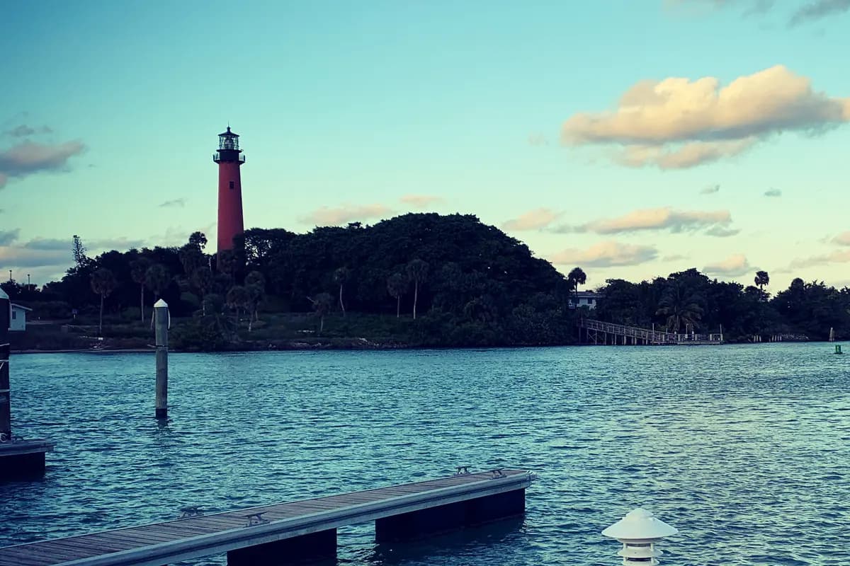 The red brick Jupiter Inlet Lighthouse rising above a wooded point across the water at dusk, with a floating dock and a white power pedestal in the foreground
