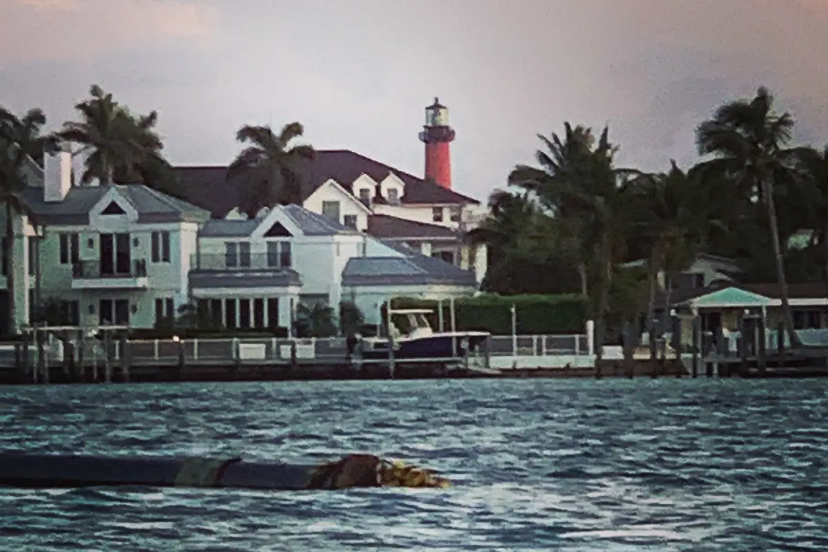 Two-story houses with docks and a moored boat on the water in Jupiter at sunset, coconut palms on either side and the red Jupiter Inlet Lighthouse rising behind the rooftops under pink clouds