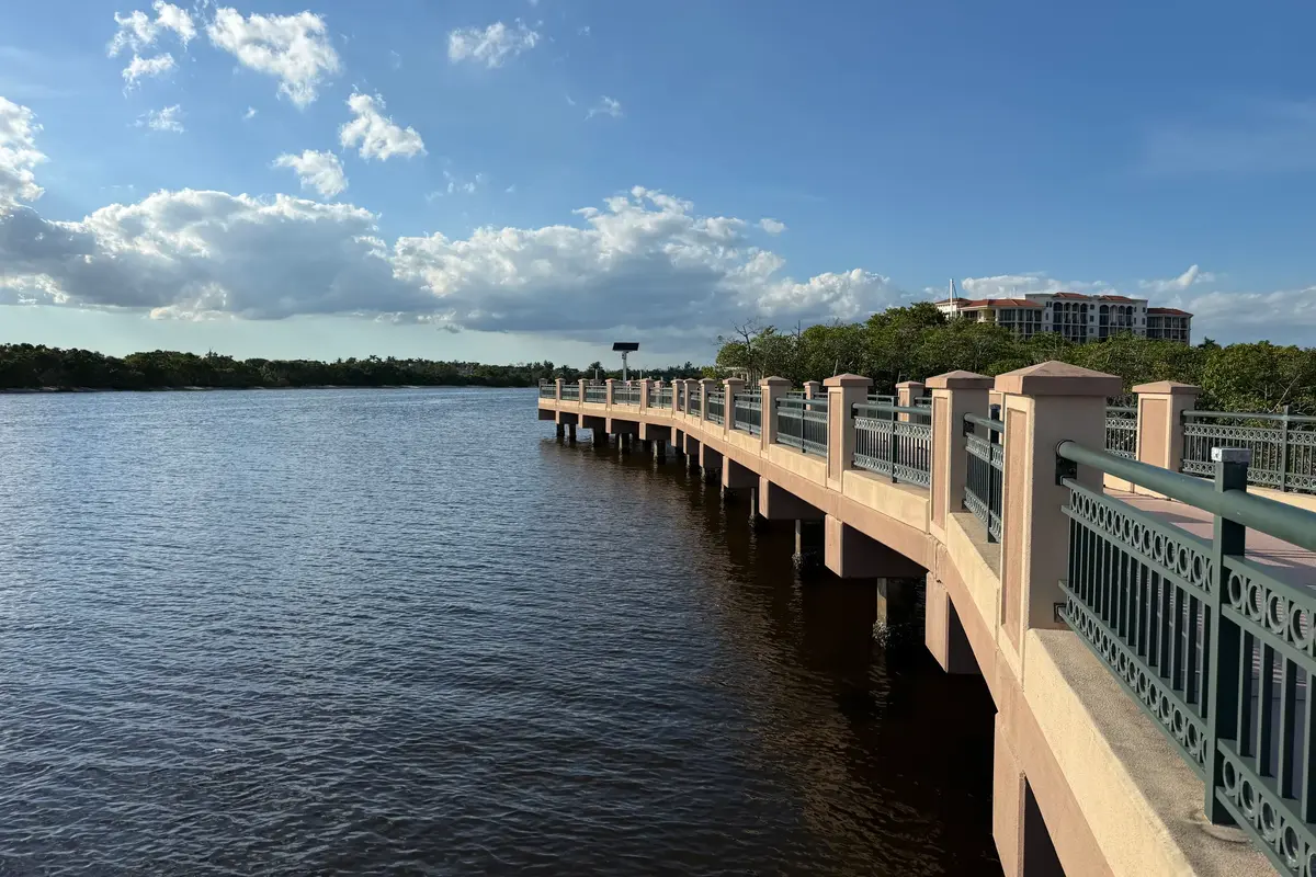 The Jupiter Riverwalk boardwalk curving out over the Intracoastal Waterway on concrete piers, with mangroves and a mid-rise condominium building behind under a blue sky with scattered clouds