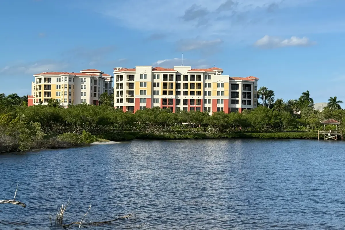 Two mid-rise condominium buildings in yellow and cream with terracotta roofs on the far shore of the Intracoastal Waterway in Jupiter, seen across the water beyond a line of mangroves under a blue sky