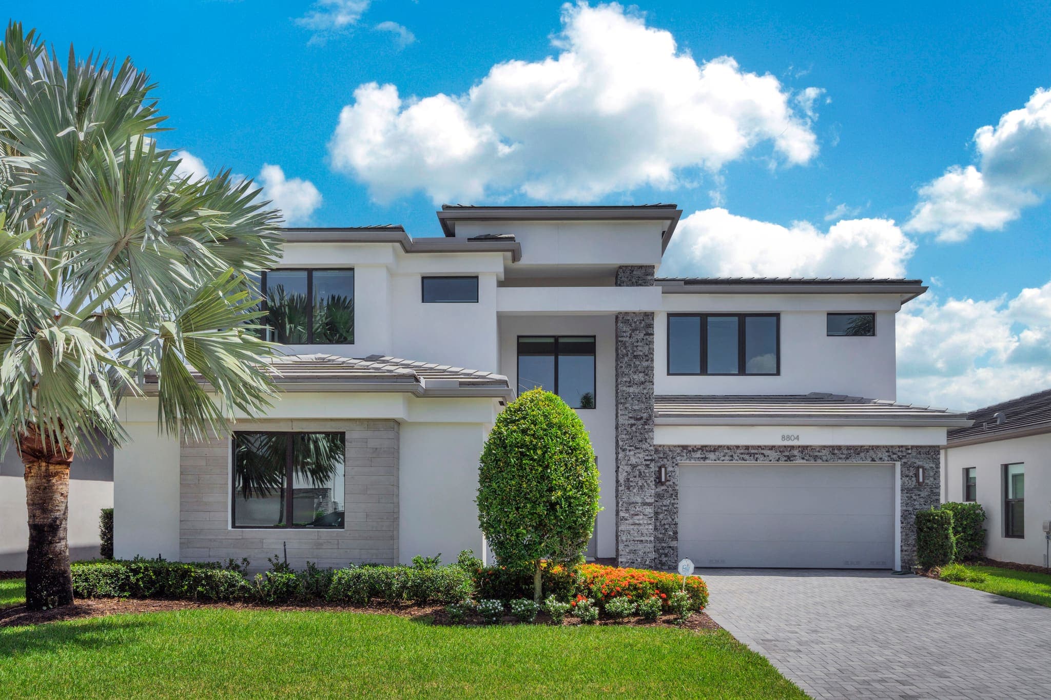 Front elevation of 8804 Skyward Street — a two-story contemporary home in white stucco with low hipped tile rooflines, black-framed windows, a stacked-stone entry column and a paver drive, framed by palms