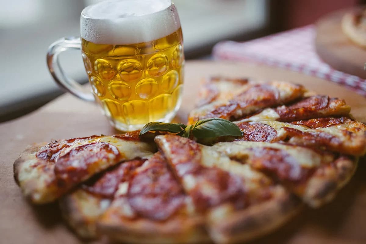 A pepperoni pizza cut into slices on a wooden board beside a frosted mug of golden beer on a rustic restaurant table, softly lit by a window