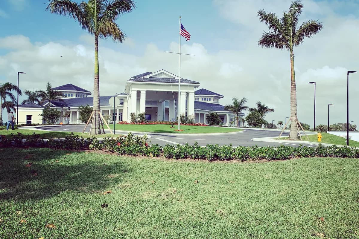 A white clubhouse with a columned entrance, a flagpole and two royal palms behind a landscaped drive