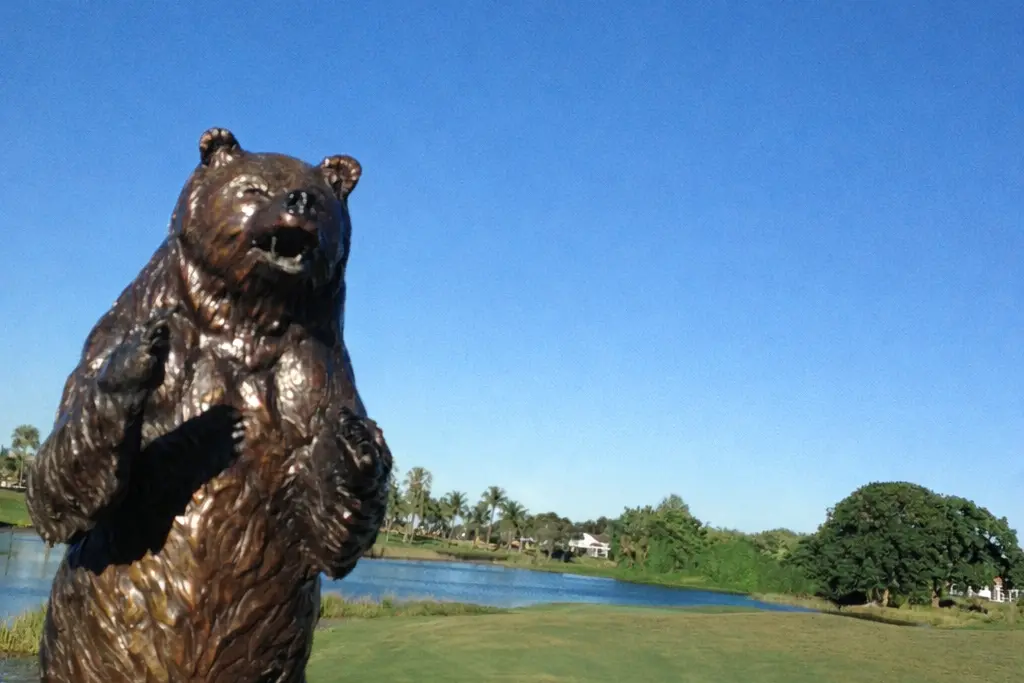 The bronze Bear Trap bear statue beside a lake and fairway on the Champion Course at PGA National in Palm Beach Gardens under a clear blue sky