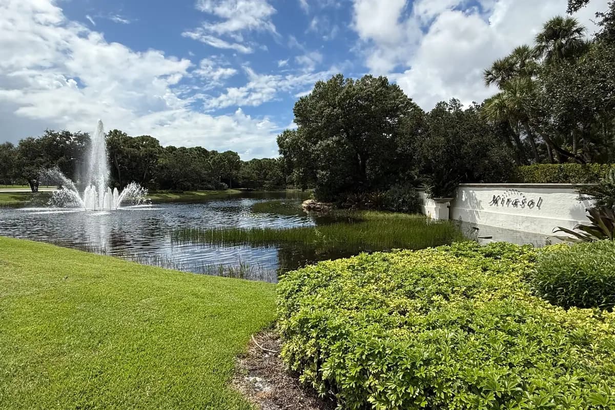 A jet fountain in a lake beside the Mirasol sign wall in Palm Beach Gardens, with clipped hedges and a lawn in the foreground and oaks and palms behind under a blue sky with scattered clouds