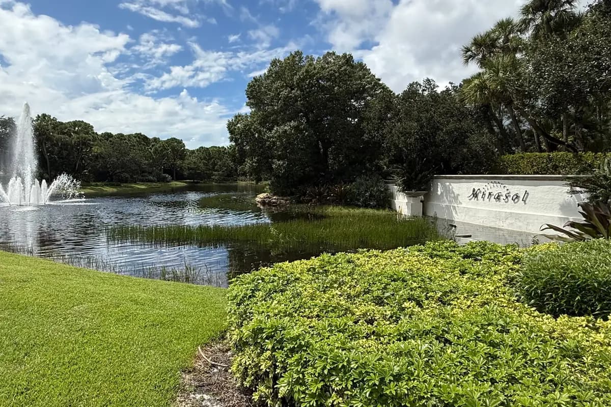 A jet fountain in a lake and the white Mirasol sign wall behind a bank of clipped yellow-green hedges, with live oaks and cabbage palms under a blue sky with scattered clouds, in Palm Beach Gardens