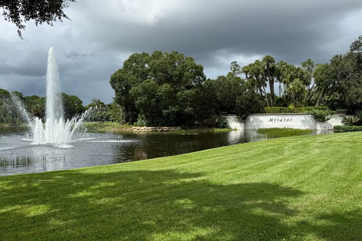 The Mirasol sign wall beside a lake with a jet fountain under a bank of dark storm clouds, with a lawn in the foreground and oaks and palms behind, in Palm Beach Gardens