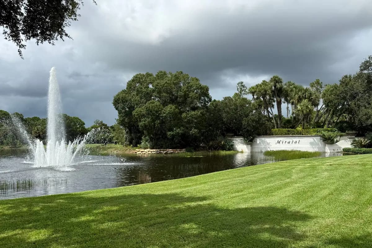 A bank of dark storm clouds over a lake with a jet fountain and the Mirasol sign wall, with live oaks and cabbage palms behind and a mown lawn in the foreground, in Palm Beach Gardens