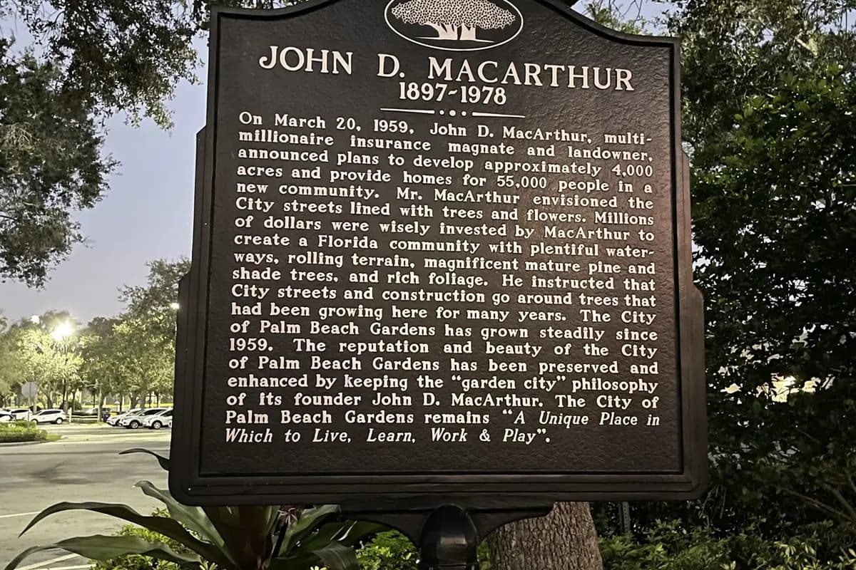A black cast-metal historical marker headed John D. MacArthur 1897 to 1978, with a banyan tree emblem and its full inscription readable, under a live oak at dusk at the Palm Beach Gardens City Hall campus