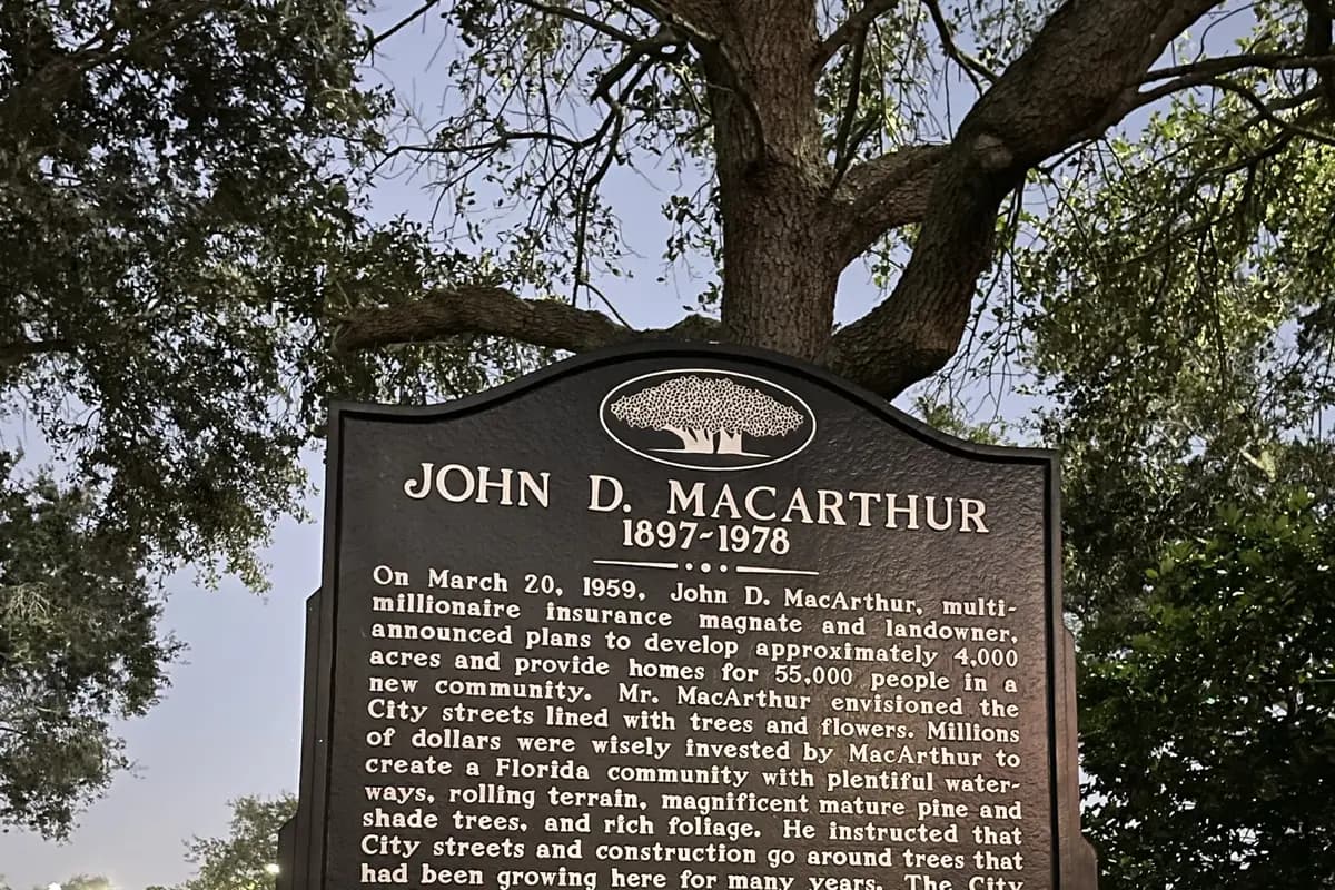 A black cast-metal historical marker headed John D. MacArthur 1897 to 1978 beneath the branches of a live oak at dusk at the City Hall campus in Palm Beach Gardens