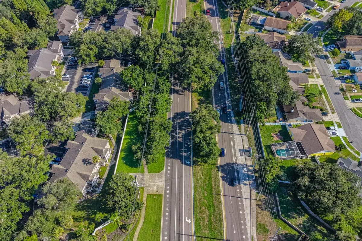 An aerial view of a divided boulevard with traffic, flanked by townhomes, pool-cage houses and cul-de-sacs under mature trees