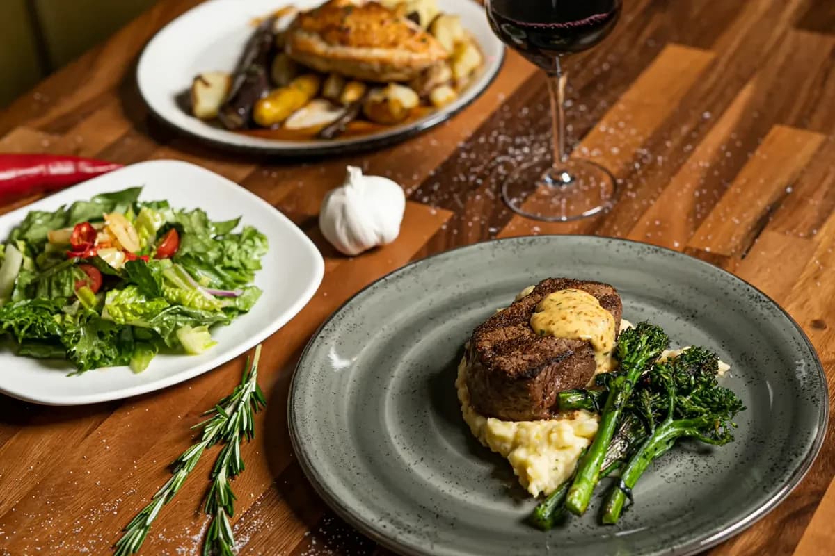 A wooden table set with a filet and mashed potatoes, a green salad, a roasted chicken dish and a glass of red wine