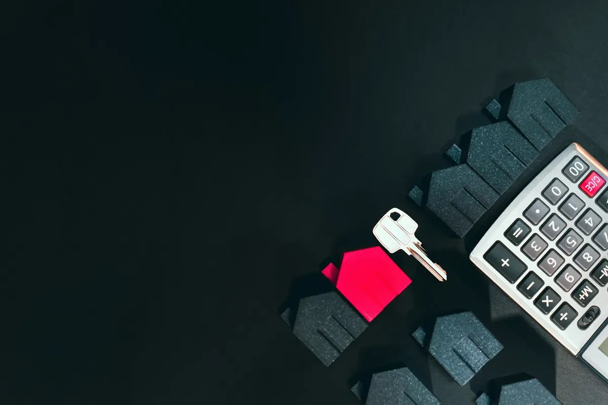 A house key, a red house model, dark house models and a calculator on a black surface