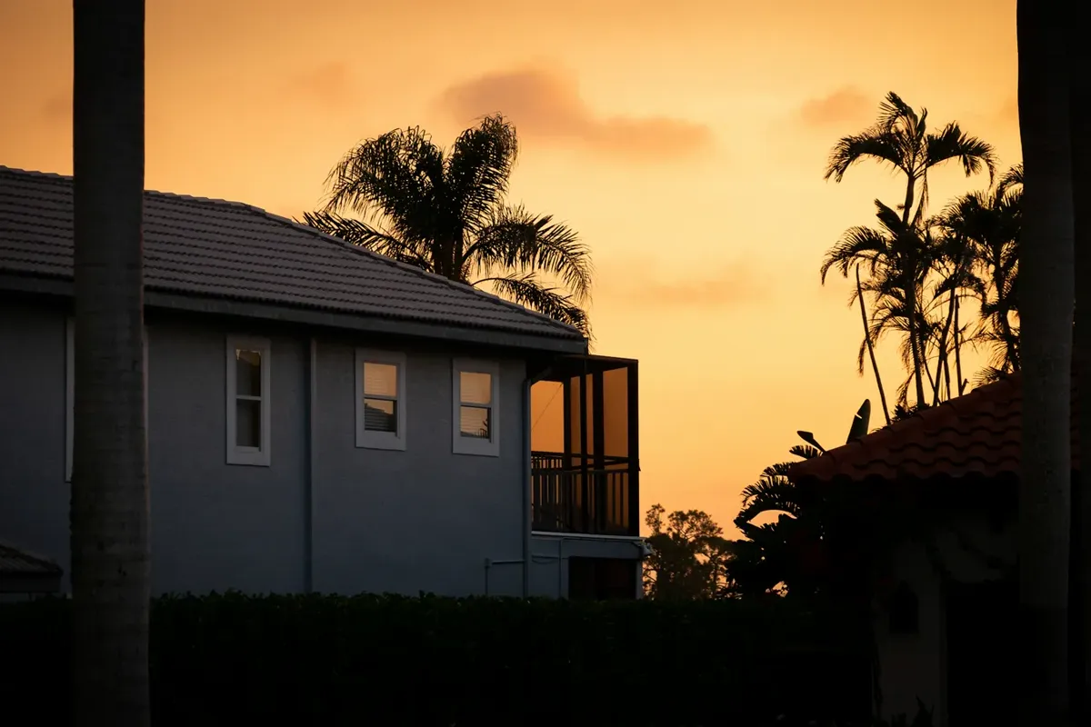 A tile-roof two-story house and palm silhouettes against an orange sunset sky