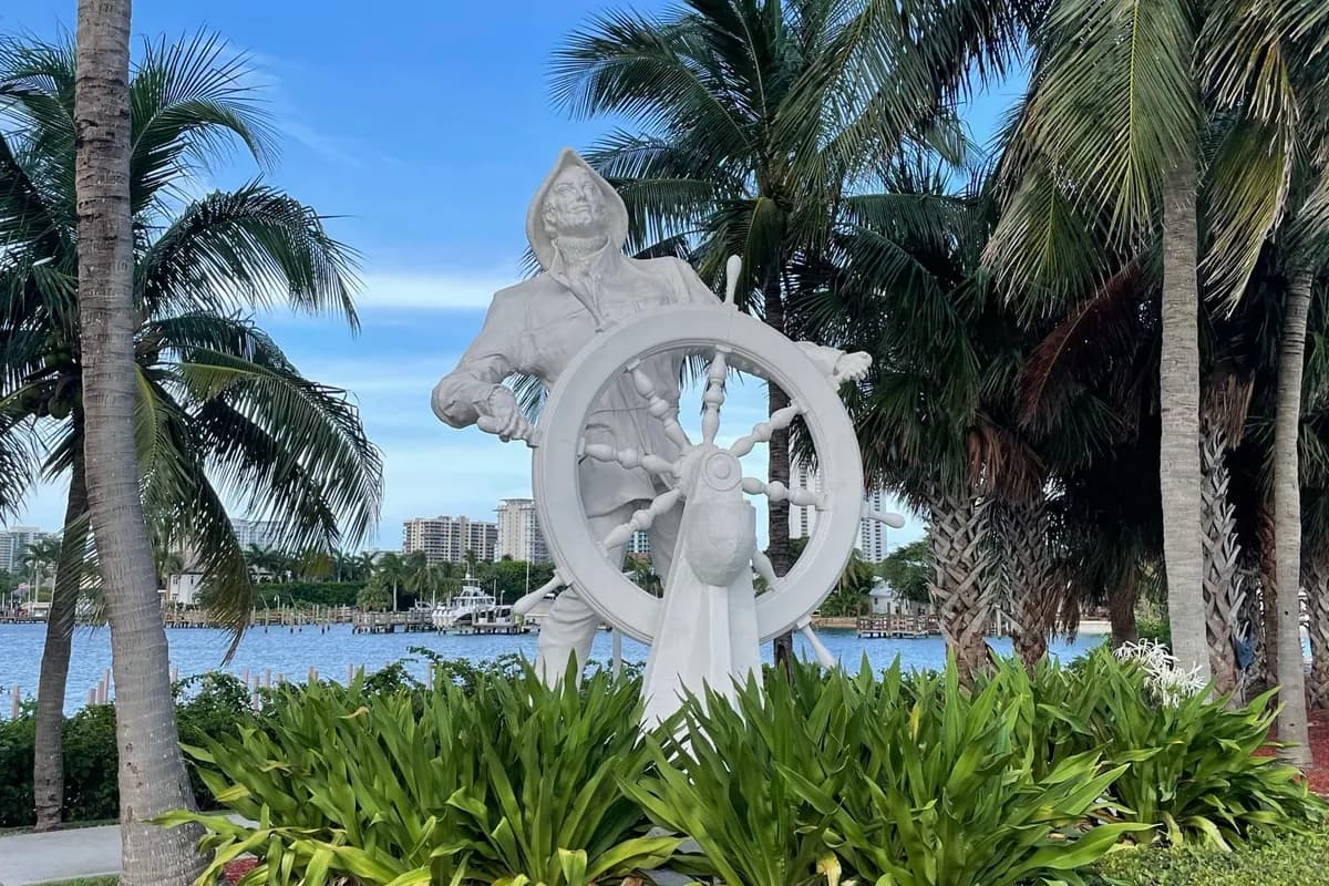 A white statue of a fisherman in a rain hat at a ship's wheel, among palms beside the Lake Worth Lagoon where the Blue Heron Bridge arrives on Singer Island