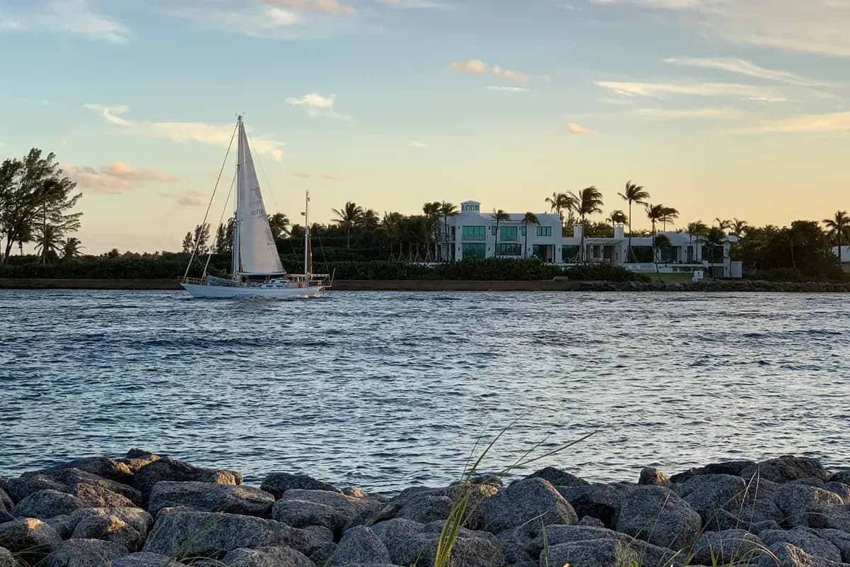A sailboat heading through the Lake Worth Inlet at sunset, seen over the rocks at the south tip of Singer Island, with Palm Beach on the far shore