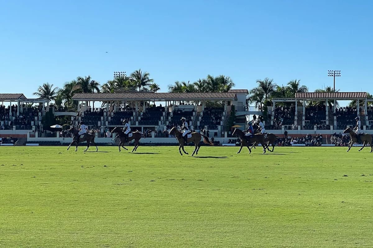 A long line of riders and horses on a grass polo field in front of a grandstand with palm trees behind