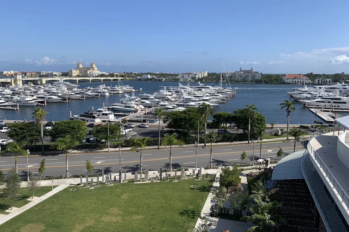 Flagler Drive and the downtown West Palm Beach waterfront seen from above: a green lawn, the waterfront road, a marina full of moored yachts on the Lake Worth Lagoon, and Palm Beach island on the far shore