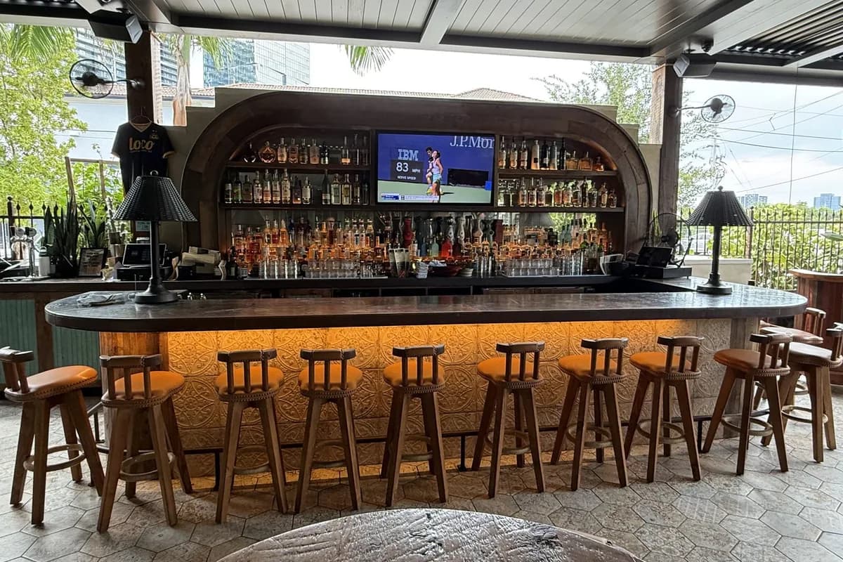 The covered patio bar at Loco Taqueria & Oyster Bar in West Palm Beach's Nora District: an arched back bar lined with bottles and a curved counter with wooden stools and amber under-lighting