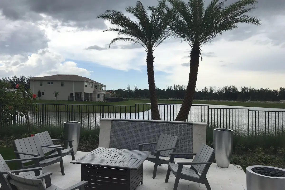 A patio with a fire table and gray outdoor chairs at a Minto model home in Westlake, with two palms, a black metal fence, a lake and a two-story house across the water under a cloudy sky