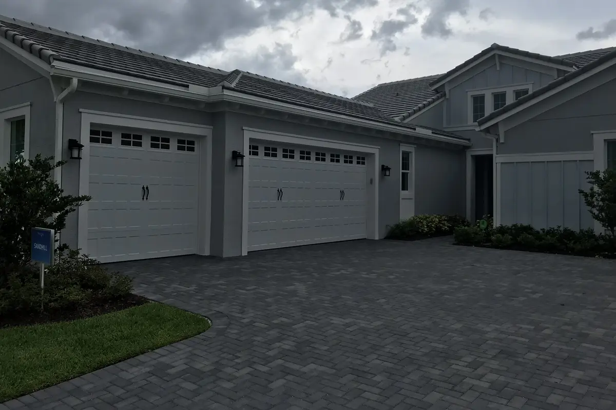 A gray single-story Minto model home in Westlake with board-and-batten siding, two white carriage-style garage doors, a wide paver driveway and a small sign in the front bed, under a cloudy sky