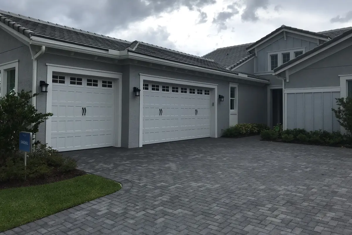 A gray single-story Minto model home in Westlake with board-and-batten siding, two white carriage-style garage doors and a paver driveway under a cloudy sky