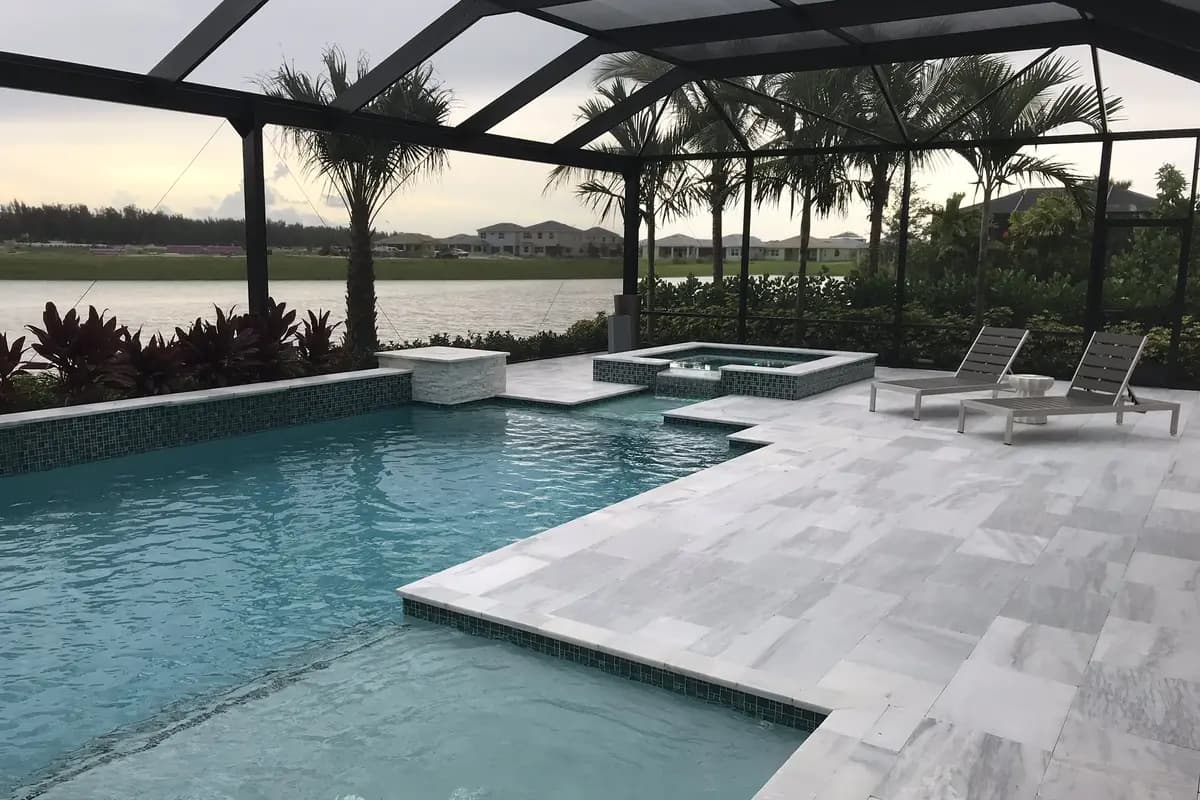 A screened pool and spa with a light stone deck and two lounge chairs at a Minto model home in Westlake, looking past palms to a lake and a row of houses under an evening sky