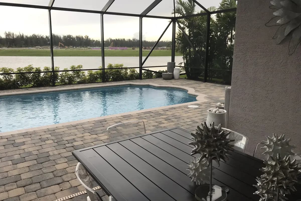 A screened lanai with a pool, paver deck and a dark outdoor table at a Minto model home in Westlake, looking across a lake to cleared land with excavators and stacked pipe