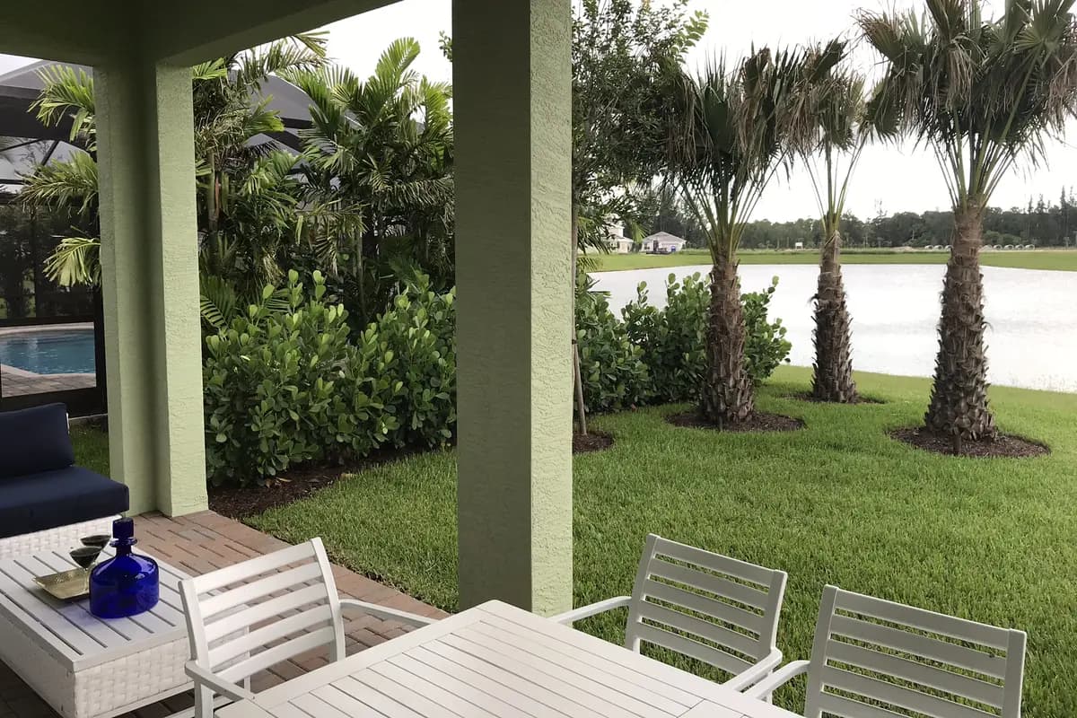 A covered patio with white outdoor furniture at a Minto model home in Westlake, opening onto a lawn with three palms and a lake, with a screened pool at left and houses across the water