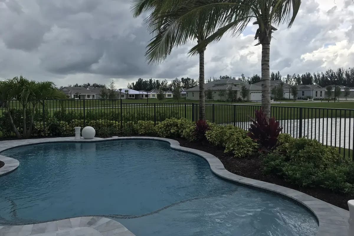A curving backyard swimming pool with a stone ball fountain and red-leafed plants at a Minto model home in Westlake, with two palms, a black metal fence, a lake and single-story homes beyond under a cloudy sky