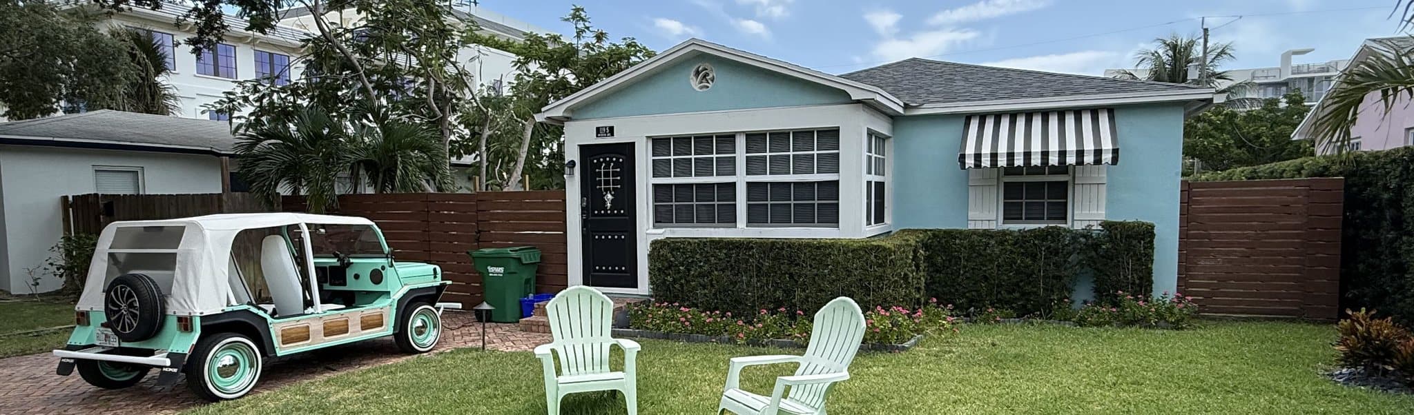 Turquoise one-story beach cottage with white trim, Adirondack chairs on the lawn and an open-sided Moke parked on the paver driveway in Delray Beach