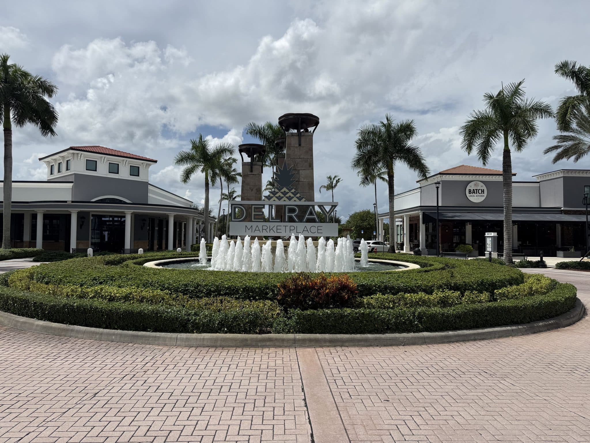 The Delray Marketplace sign on its stone tower above the entrance fountain, with palms and storefronts behind
