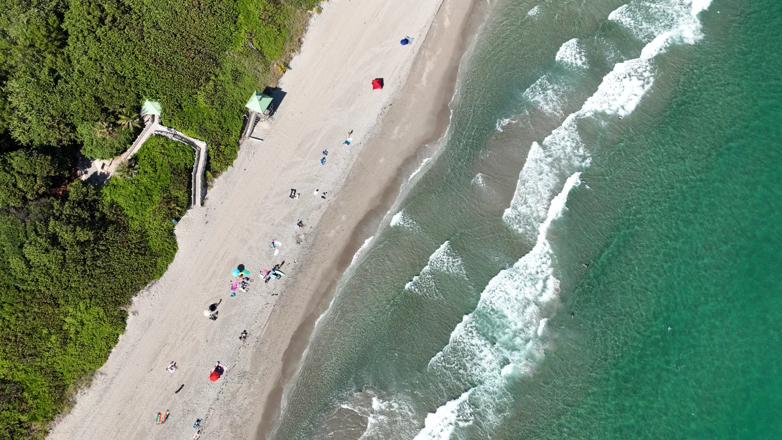 Top-down drone view of a sandy beach with turquoise surf and dune-vegetation boardwalk