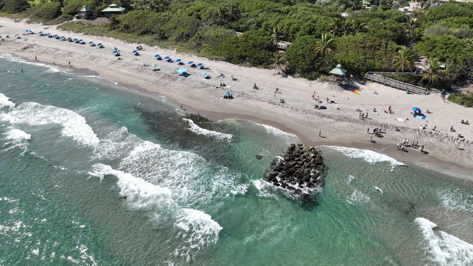Aerial view of a Florida beach with turquoise surf, rock outcrop, and beachgoers under blue umbrellas